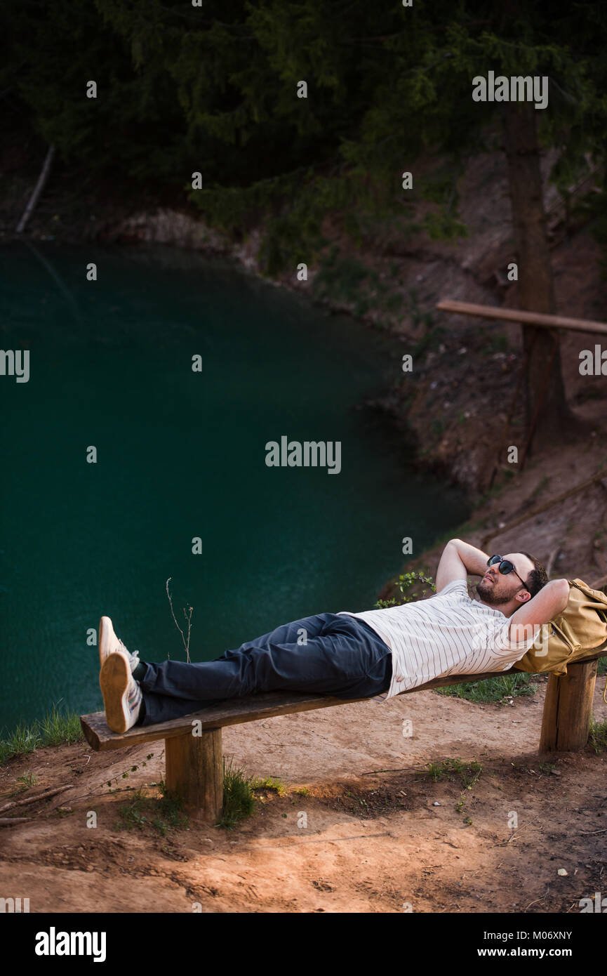 Mann draußen entspannen. Tragen ein weißes T-Hemd, dunkle Hose, Sonnenbrille, ist ein junger Mann auf Holzbank gegen See in einem Wald im freien liegend Stockfoto