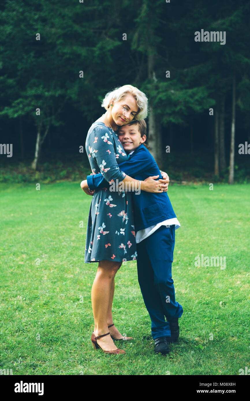 Lächelnde Mutter und Sohn umarmen vor einem Wald auf einer Wiese im Sommer. Stockfoto