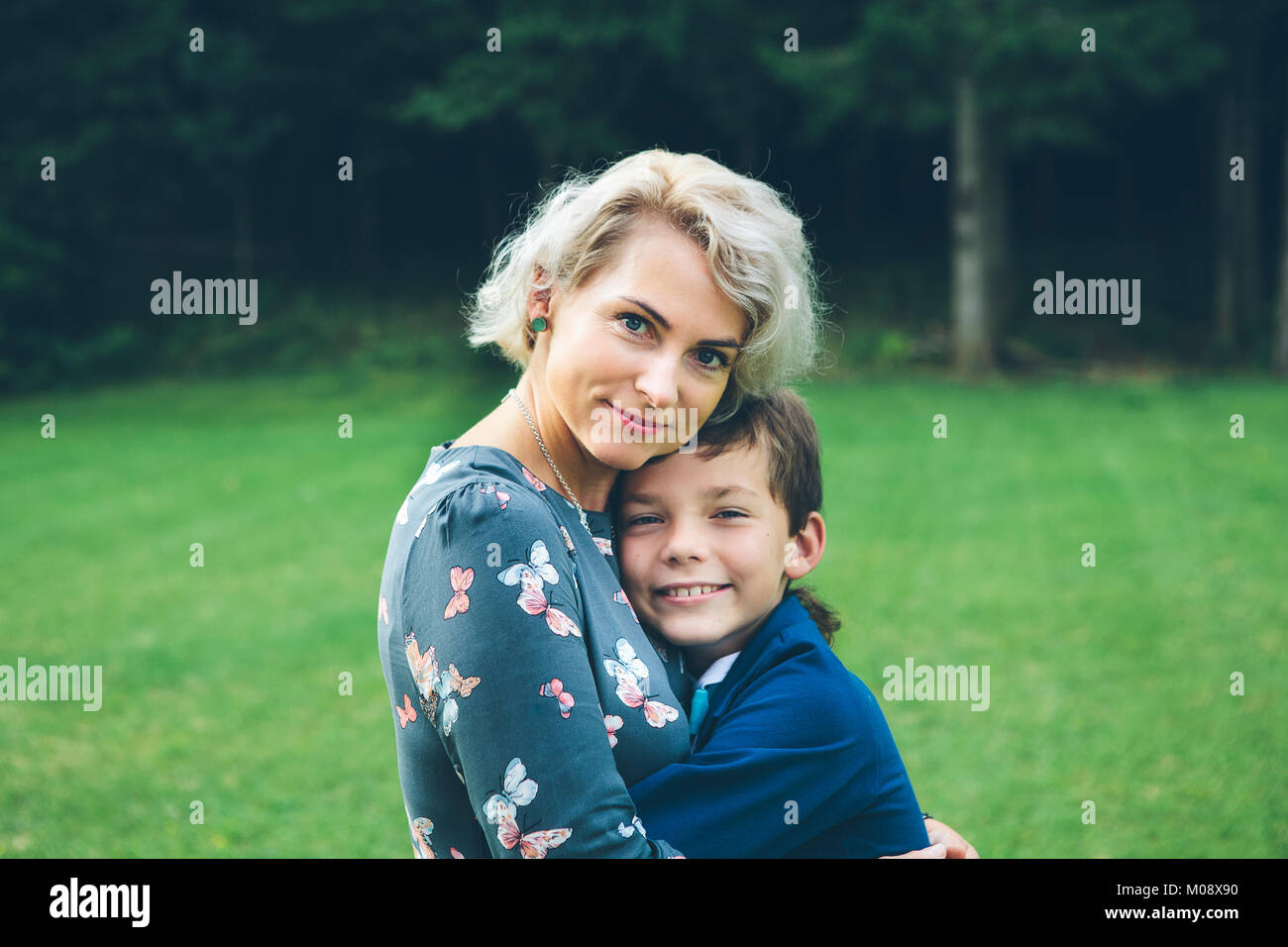 Lächelnde Mutter und Sohn umarmen vor einem Wald auf einer Wiese im Sommer. Stockfoto