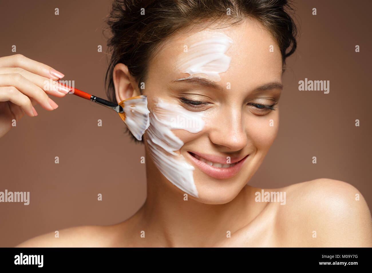 Junge Frau mit feuchtigkeitsspendenden Gesichtsmaske und Hände der Kosmetikerin auf braunem Hintergrund. Foto der hübschen Frau erhält die Behandlungen im Spa. Beauty & Stockfoto