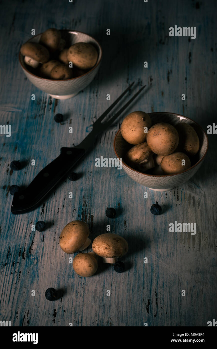 Vertikale Foto mit essbaren Pilzen. Pilze mit braunen Kappen in zwei Schüsseln und auf Vintage blau Holzbrett. Große Gabel zwischen Schalen gelegt. Trockene blau Stockfoto