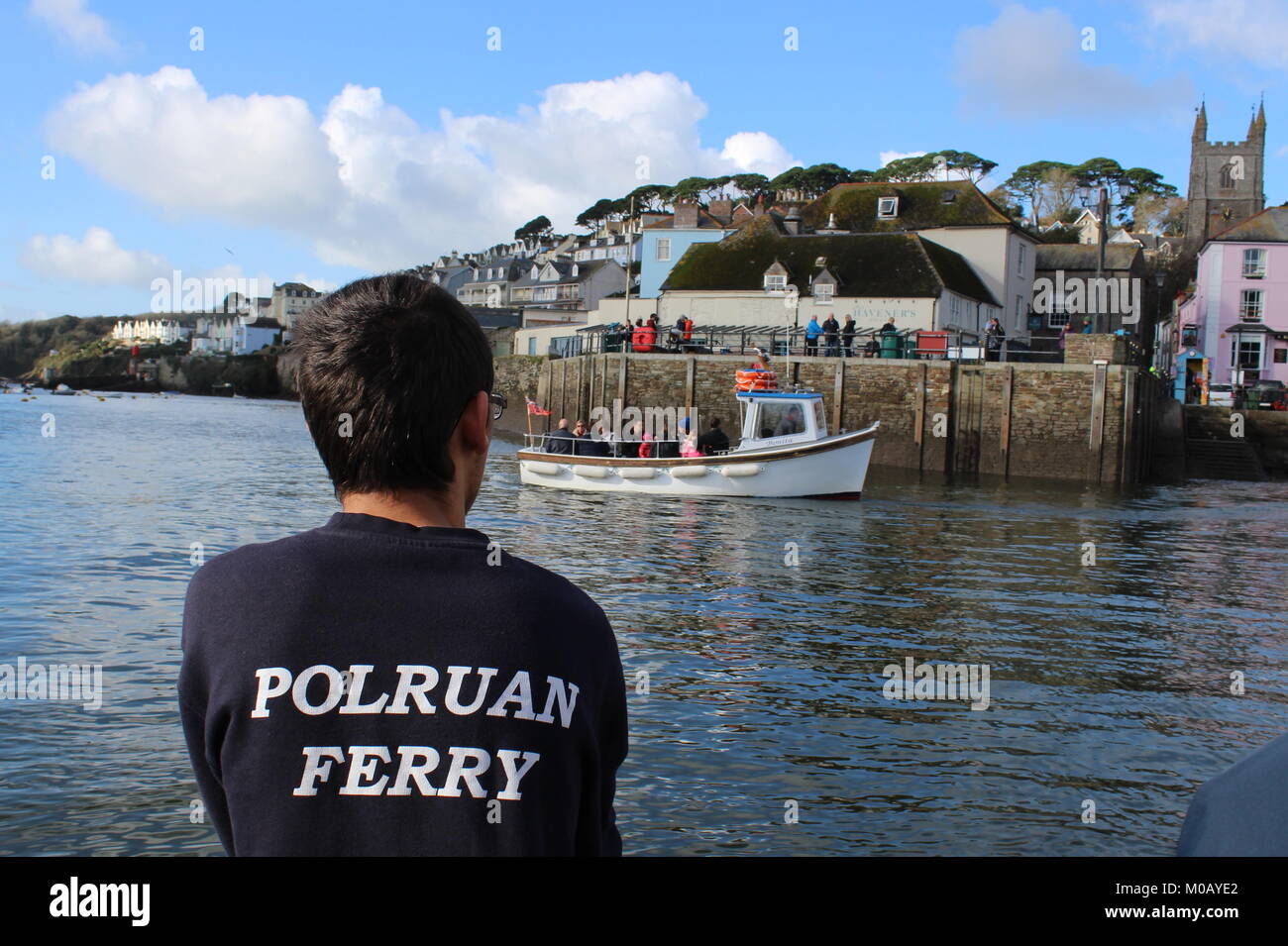 Die Rückseite der Fährmann, der ein schwarzes T-Shirt mit Polruan geschrieben als die Fähre Fowey Harbour und einem kleinen weißen Boot Kreuze vor erreicht. Stockfoto