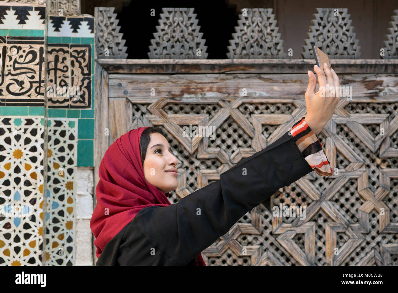 Arabische Frau in traditioneller Kleidung unter selfie mit Handy vor der Wand mit Text aus dem Koran Stockfoto