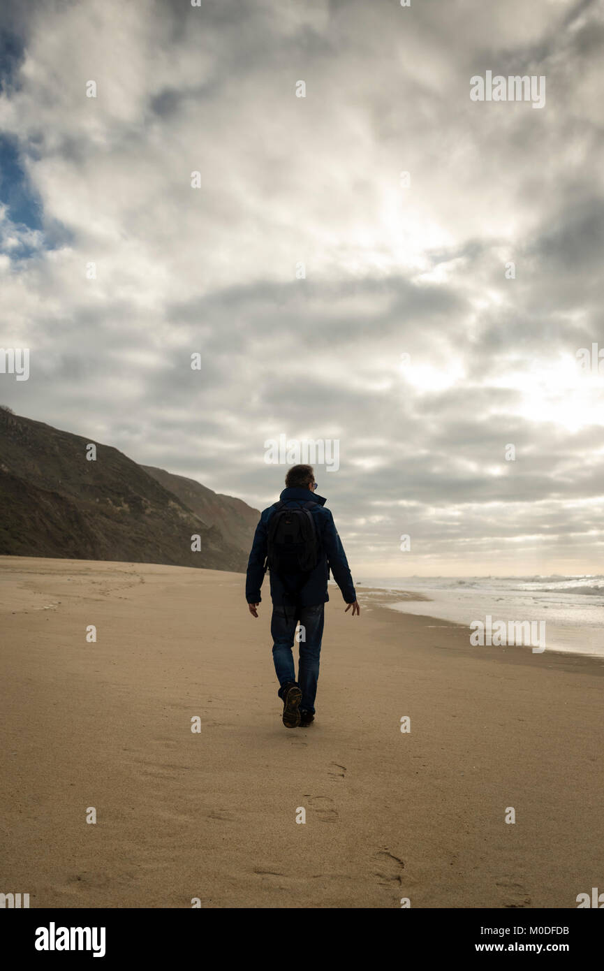 Mann in Silhouette entlang einem abgelegenen Strand trägt einen Rucksack, Moody Himmel. Stockfoto