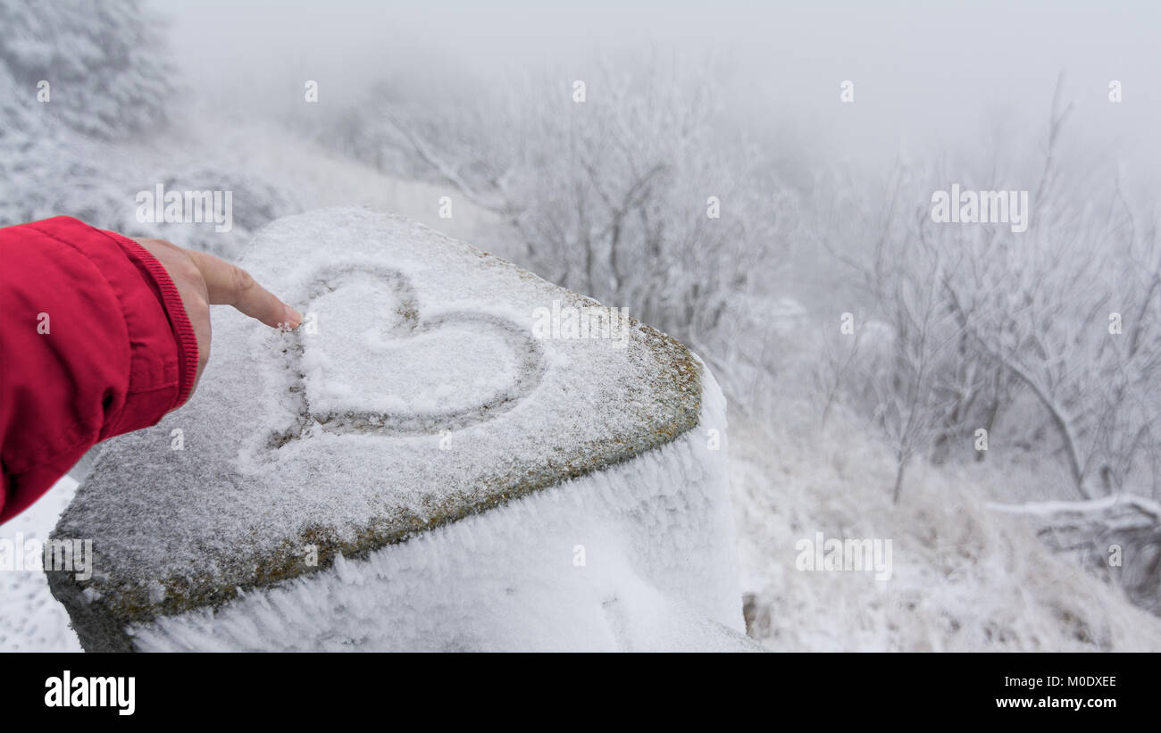 Weibliche finger zeichnet einen symbolischen Herzen für gutes Glück im Schnee. Hand in rote Hülse und Symbol der Liebe mit frostigen Winter Landschaft im Hintergrund. Stockfoto