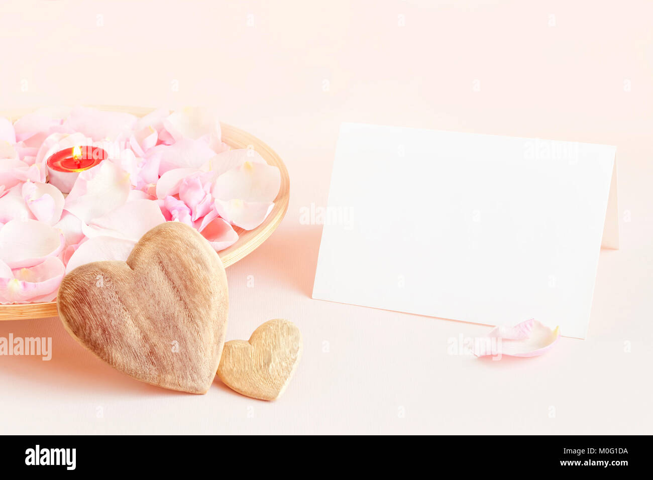 Leere weiße Karte auf rosa Hintergrund mit Holzplatte gefüllt mit rosa Rosenblätter und Kerzen und zwei hölzerne Herz für St Valentines Tag Stockfoto