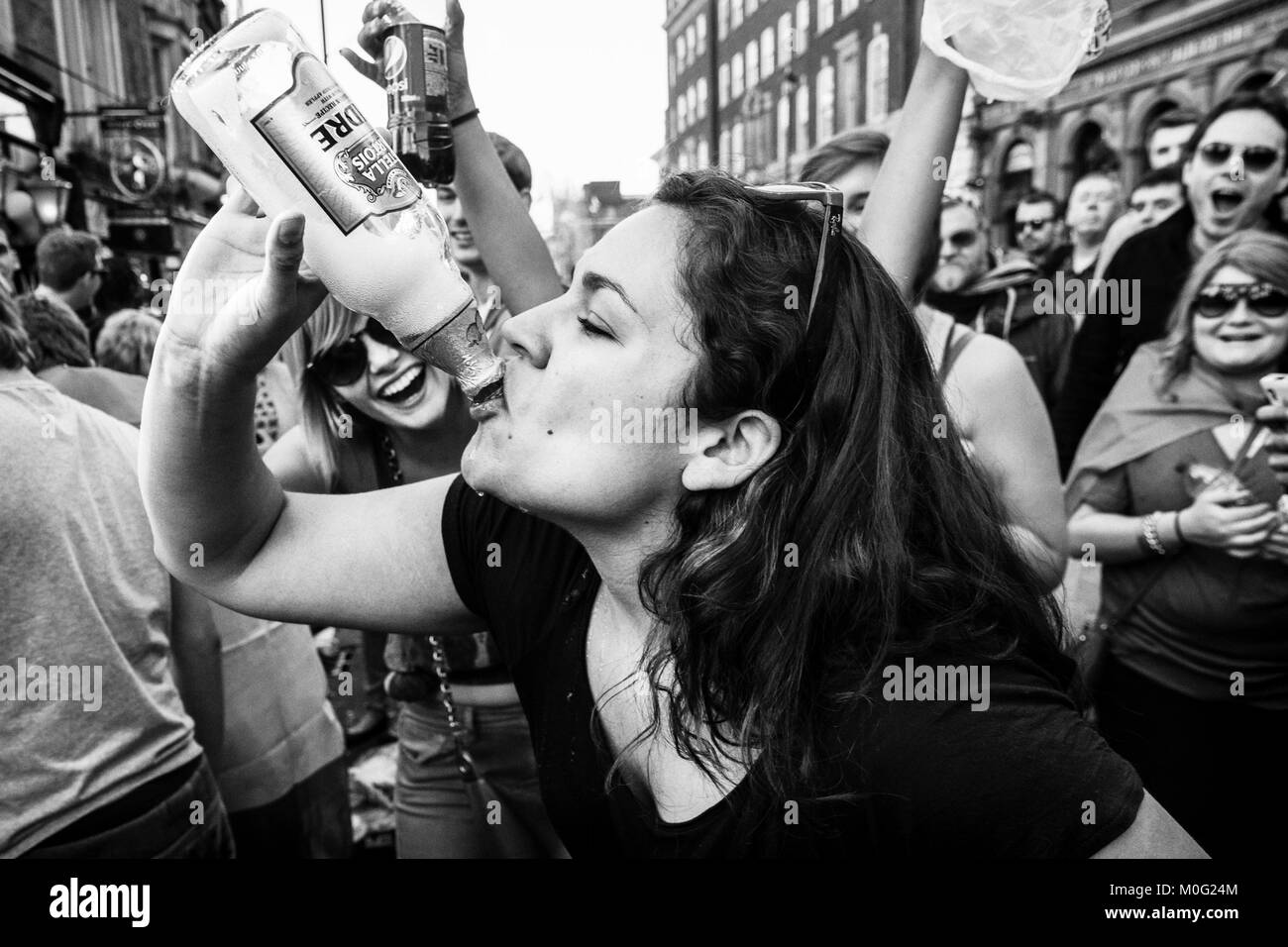 Schwarz-weiße Straßenfotografie in London: Junge Frau wird von der Menge angefeuert, als sie bei der Straßenparty aus der Flasche trinkt. Stockfoto