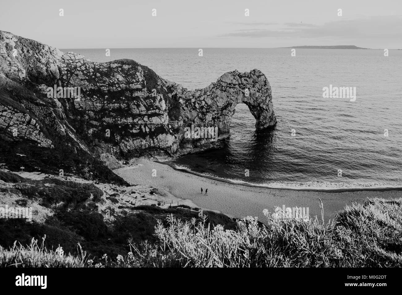 Schwarz & Weiß Blick vom Hügel über Menschen zu Fuß auf einem Strand am Meer und Durdle Door, ein natürlicher Kalkstein Bogen auf Jurassic Küste, UK. Stockfoto