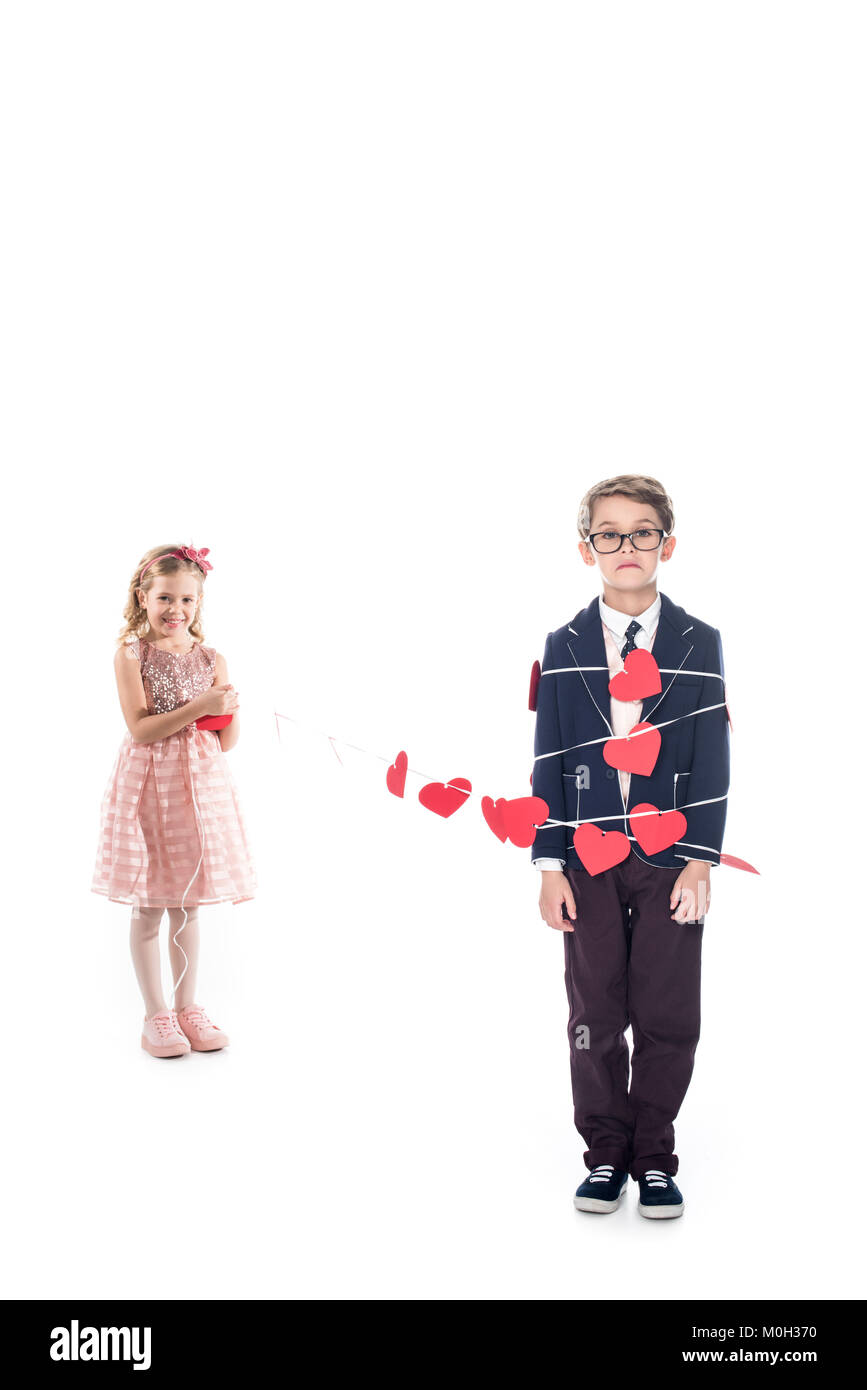 Bezaubernd lächelnde Mädchen binden Stilvolle Junge mit Seil und roten Herzen isoliert auf weißem Stockfoto