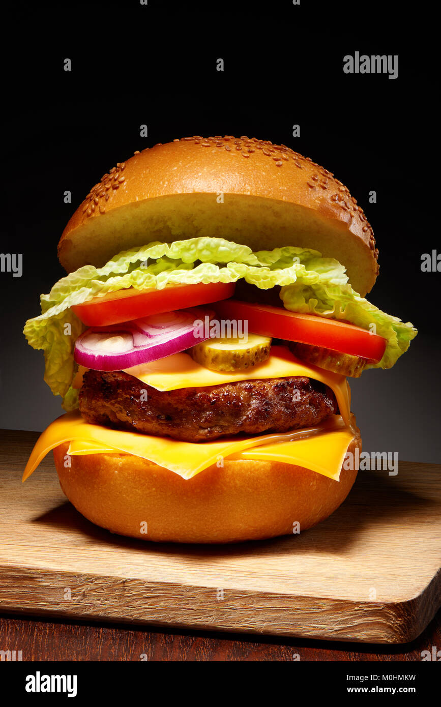 Lecker Hamburger mit Käse, Gurken, Tomaten, Zwiebeln und Salat auf Holz Schneidebrett auf dunklen Tabelle Stockfoto