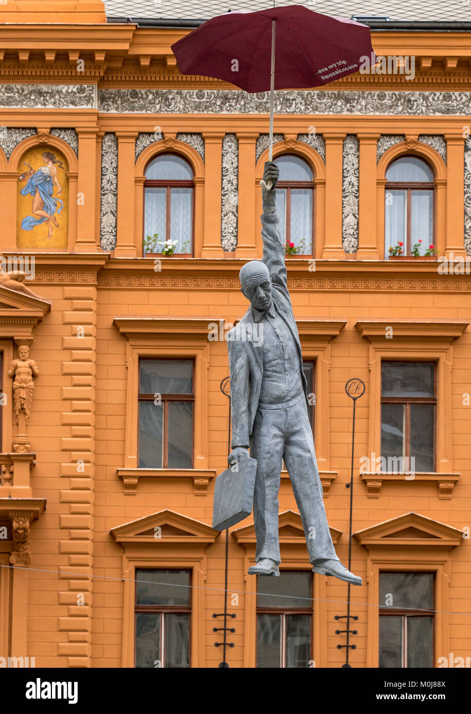 Die Umbrella Man von Prag, gegen ein orangefarbenes Gebäude gesehen, eine moderne Kunst Statue eines Mannes hängt von einem Dach hoch über einem Prager Straße ausgesetzt Stockfoto
