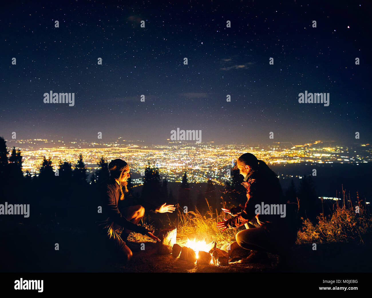 Glückliches Paar Wanderer wärmen Sie die Hände in der Nähe von Lagerfeuer unter Sternenhimmel mit Sterne und die Lichter der Stadt im Hintergrund Stockfoto