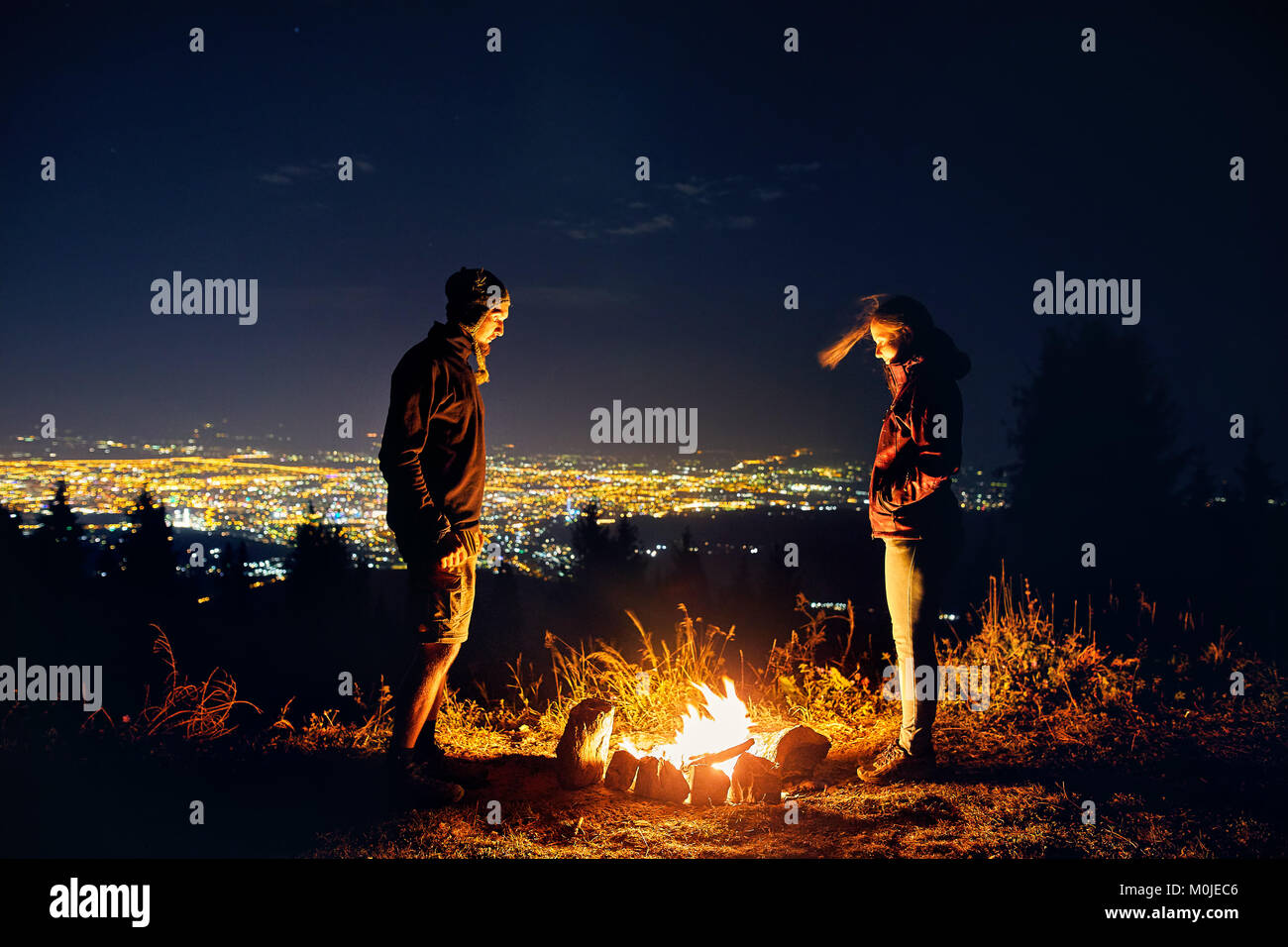 Glückliches Paar Wanderer in der Nähe von Lagerfeuer unter Sternenhimmel und die Lichter der Stadt im Hintergrund Stockfoto