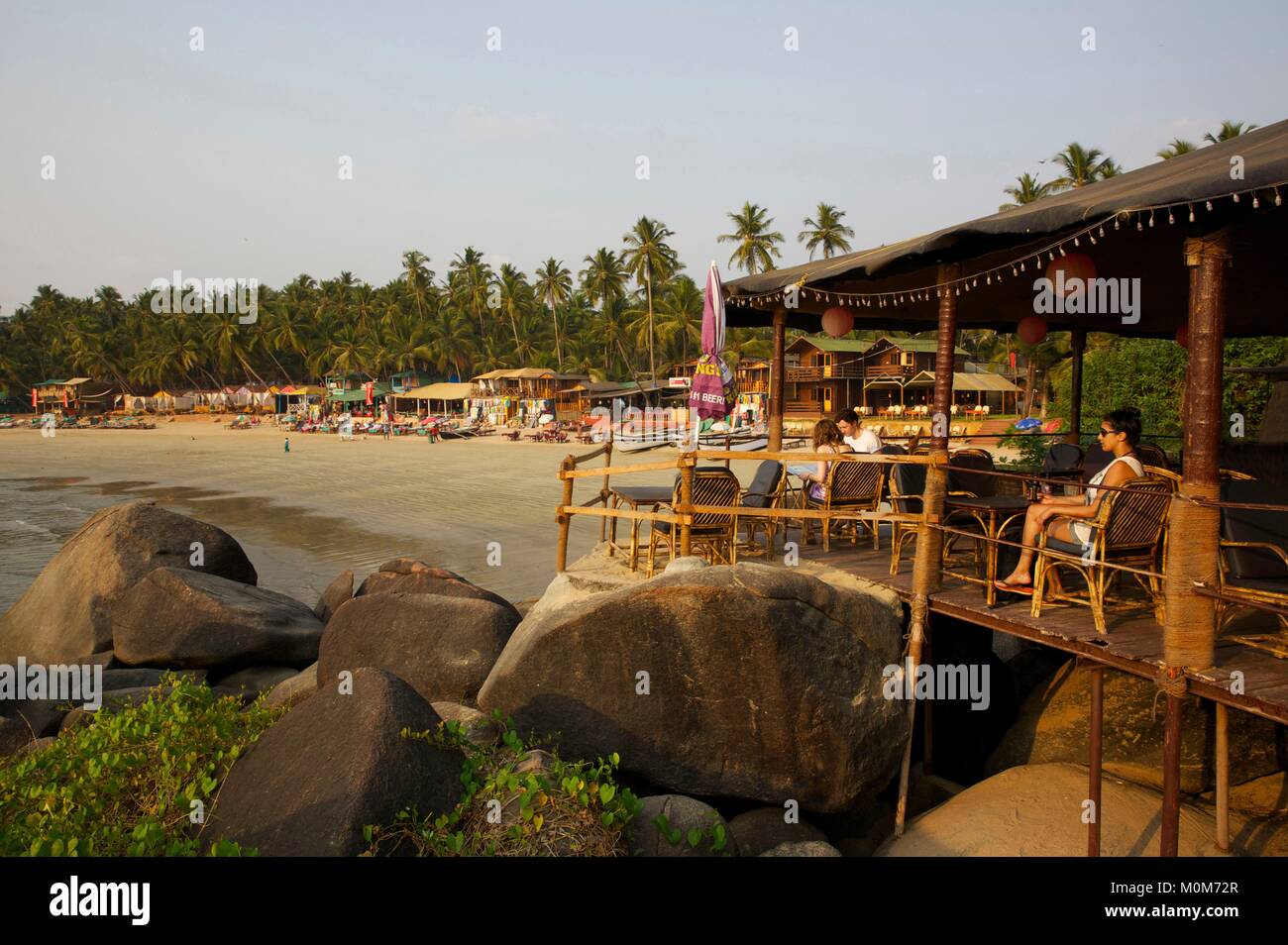 Indien, Goa, Palolem, Touristen auf der Terrasse einer Bar mit Blick auf den goldenen Sandstrand mit Restaurants und Geschäften gesäumt, zwischen Felsen und Coconut Bäume Stockfoto