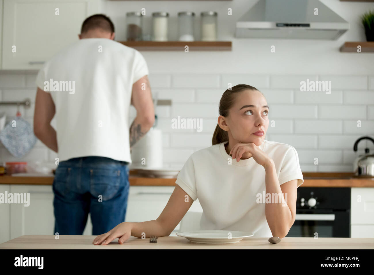 Melancholisch nachdenkliche Frau sitzt am Küchentisch, während Mann kochen Stockfoto