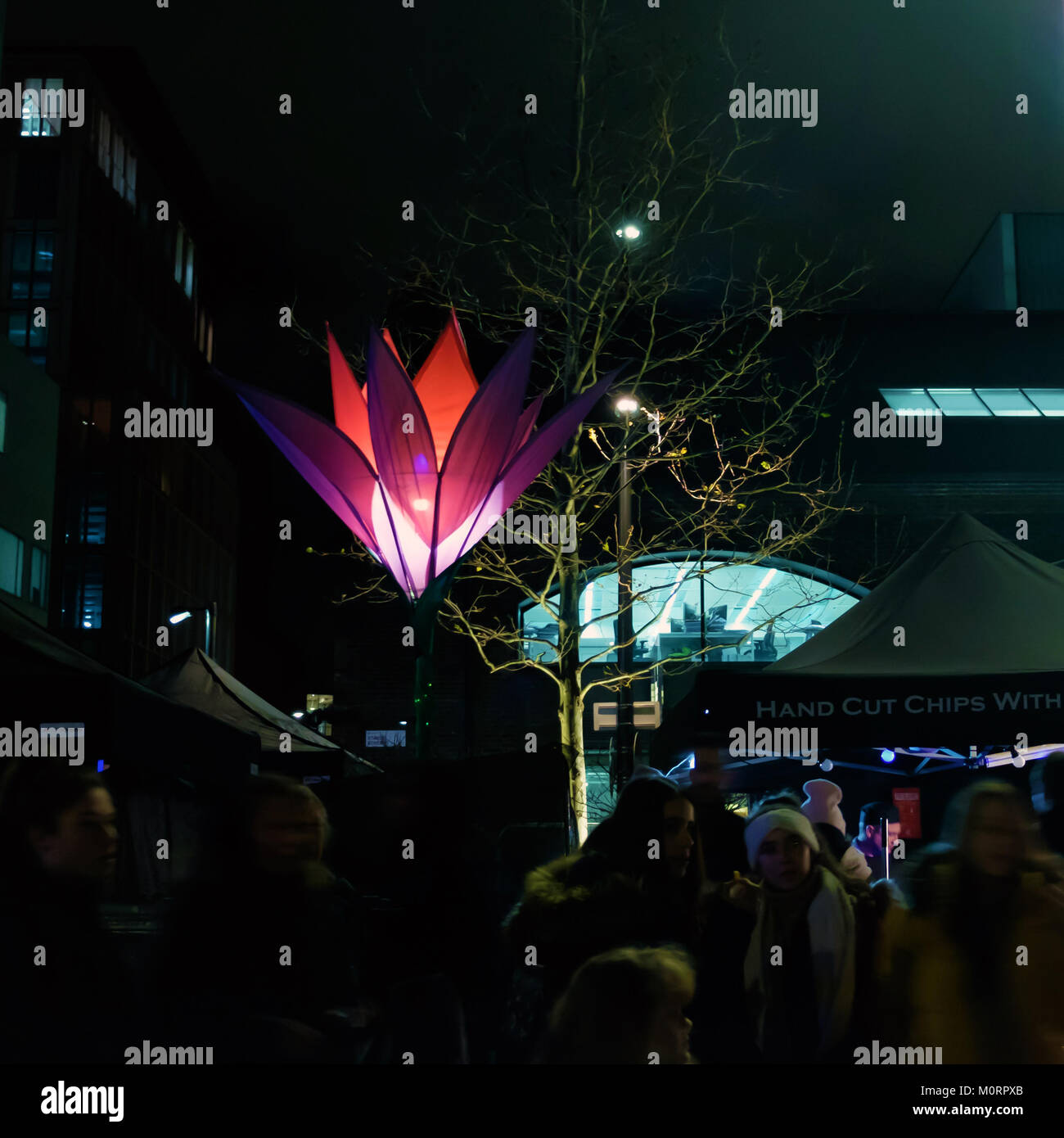 Eine grosse Neon Blume in der Nacht und Imbissstände mit Menschen kaufen Lebensmittel und Essen. Stockfoto