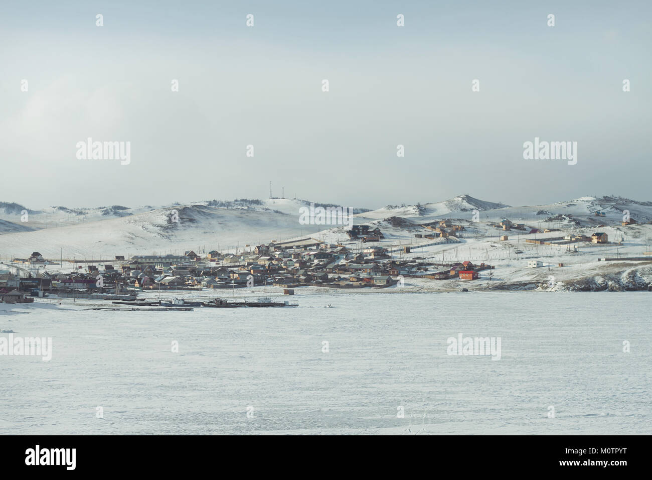 Eine ländliche Landschaft mit Häusern im Winter am Ufer des Baikalsees, die Schiffe auf dem Eis im Vordergrund auf einem schneebedeckten Bergen im Hintergrund. Stockfoto
