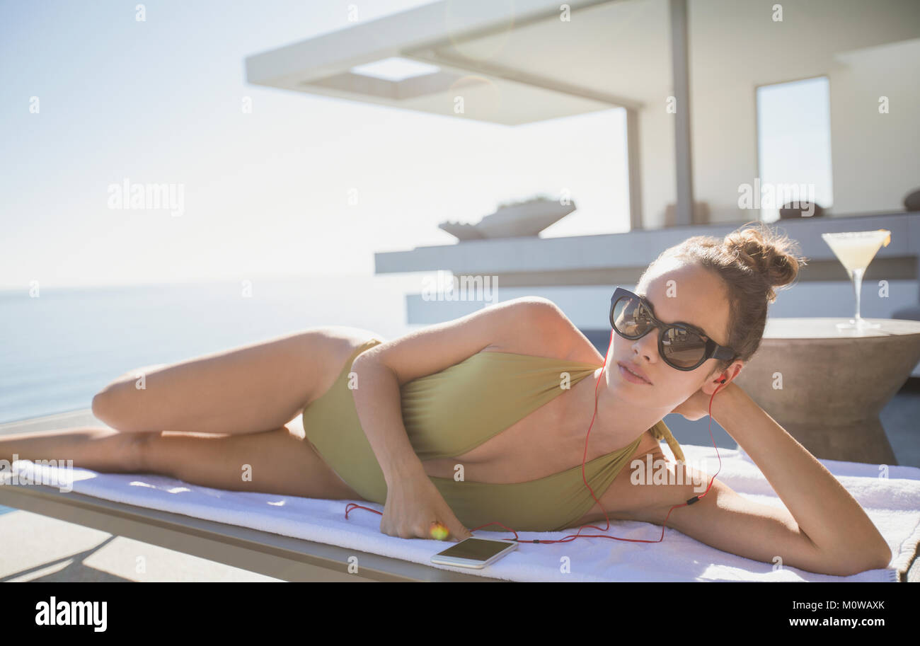 Portrait selbstbewusste Frau im Badeanzug Sonnenbaden an sonnigen Luxus Terrasse, das Hören von Musik mit smart phone Stockfoto