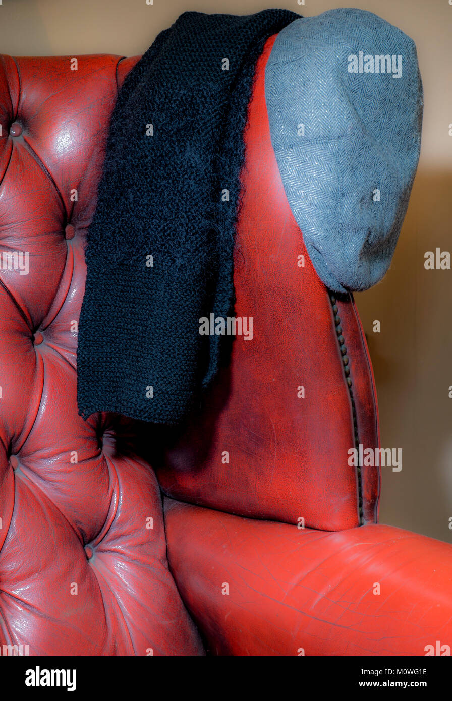 Warme Winterkleidung durch ein schwarzer Strickschal hängen an eine alte traditionelle leder ohrensessel mit einem grauen flacher Deckel neben. England, UK. Stockfoto