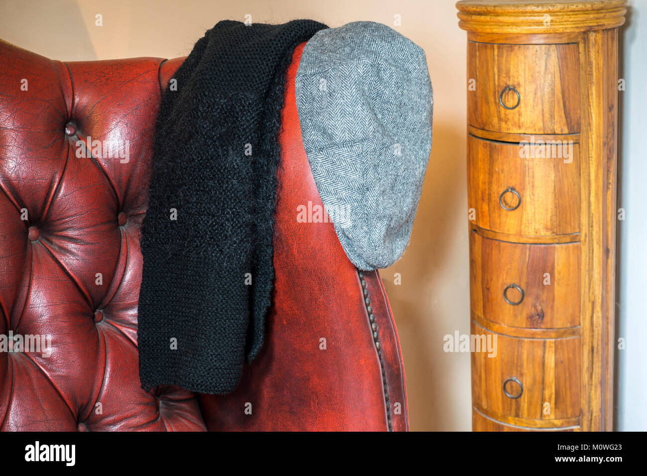 Ein schwarzer Strickschal hängen an eine alte traditionelle leder ohrensessel mit einem grauen flacher Deckel neben, und eine geschwungene Schubfach. England, UK. Stockfoto