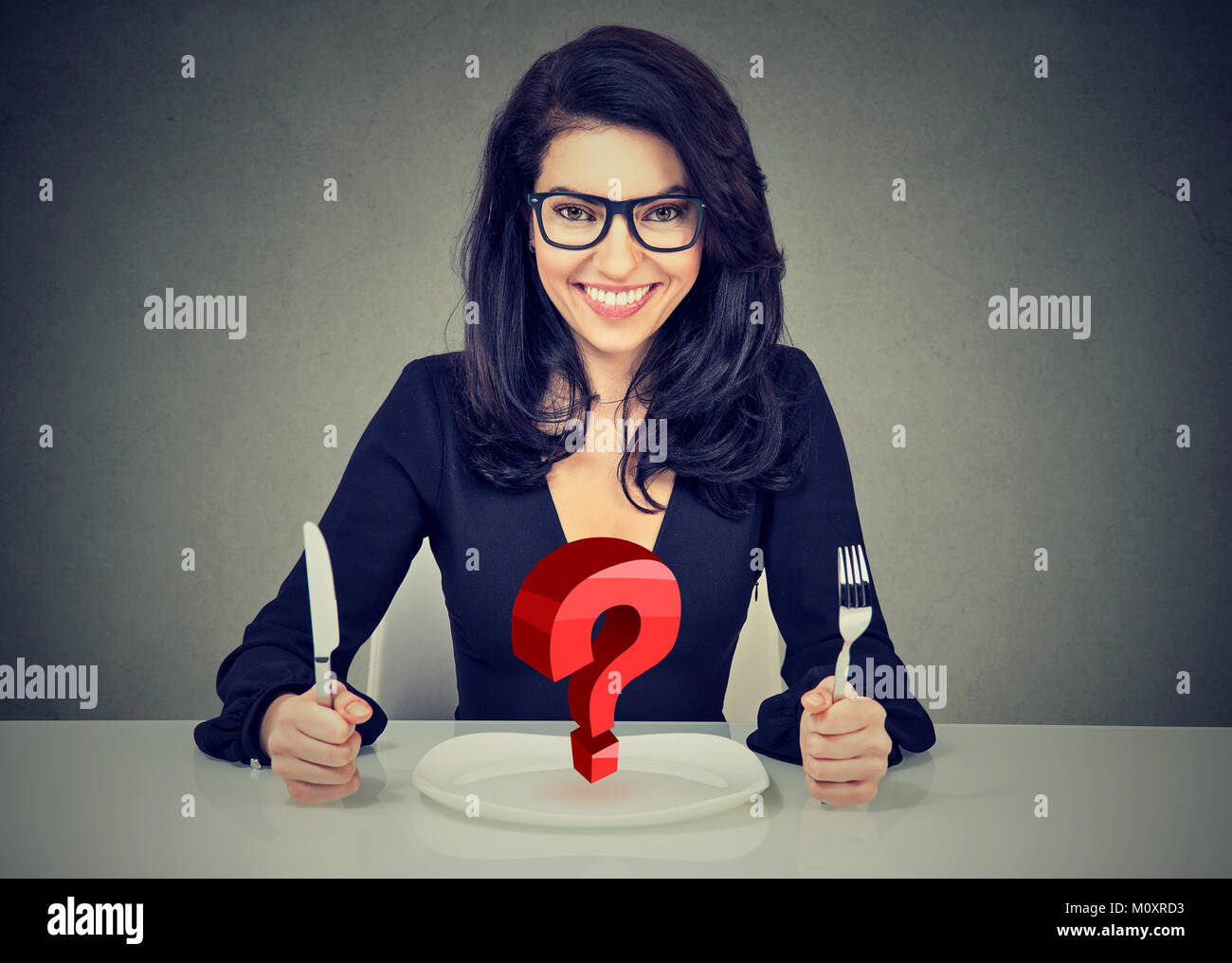 Glückliche Frau in Gläsern mit Messer und Gabel sitzen am Tisch mit leeren Teller und Fragezeichen auf grauem Hintergrund Stockfoto