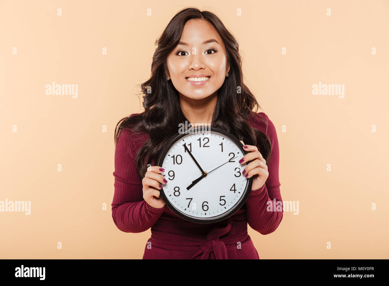 Glückliche Gefühle der asiatische Frau mit dem lockigen langen Haare, die Uhr zeigt fast 8, warten auf etwas Angenehmes über Pfirsich Hintergrund Stockfoto