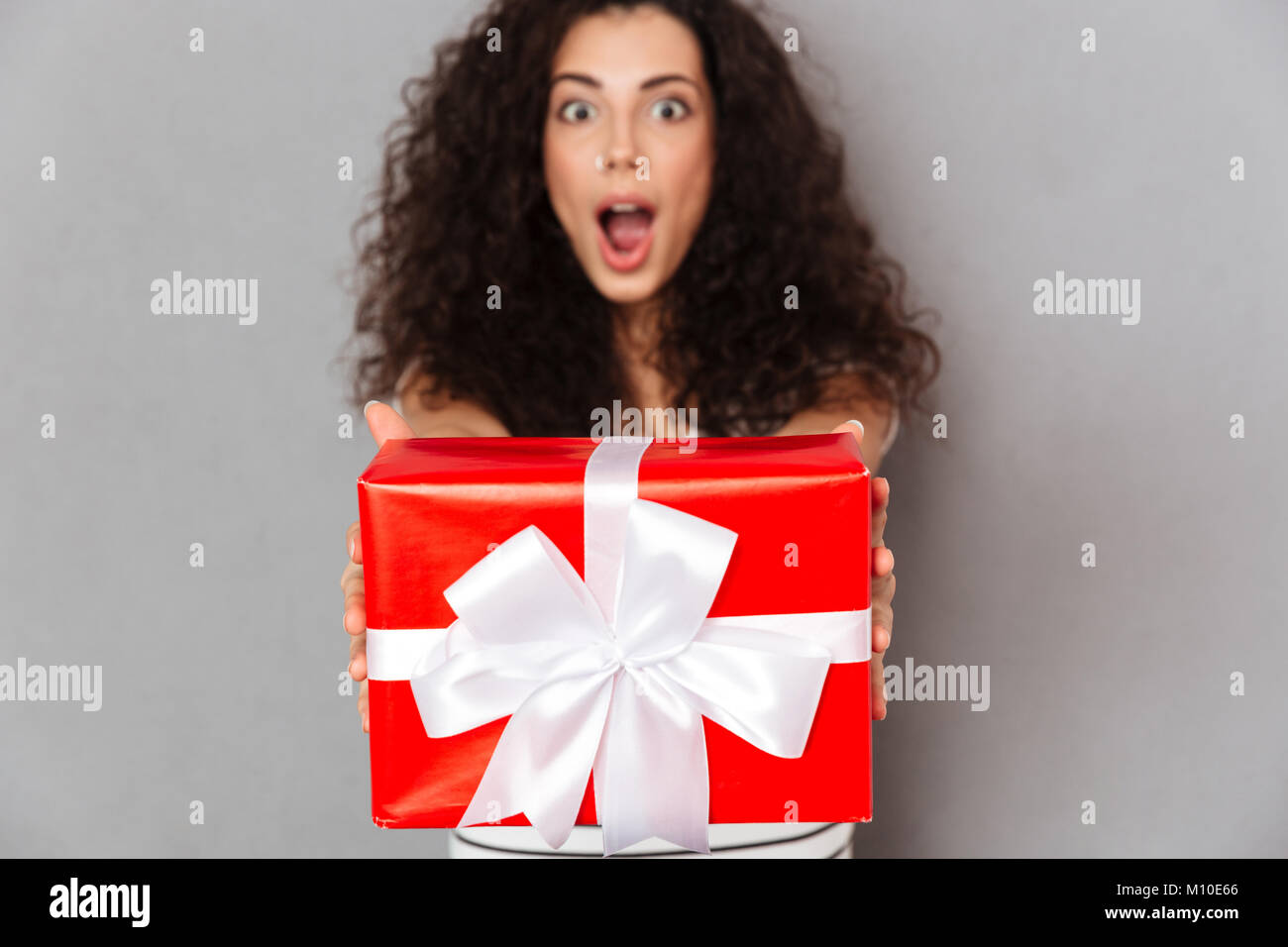 Brünette Frau in Defokussieren gegen graue Wand holding red box Geschenk - Posing mit weißen Bogen gewickelt, es mit der Kamera Stockfoto