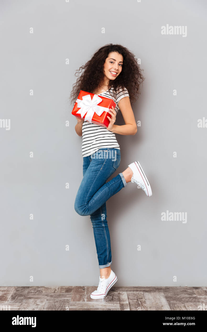 Das Bild der hübschen erwachsenen Girl posiert vor der Kamera mit Geschenk verpackt in ihren Händen, über graue Wand isoliert Stockfoto
