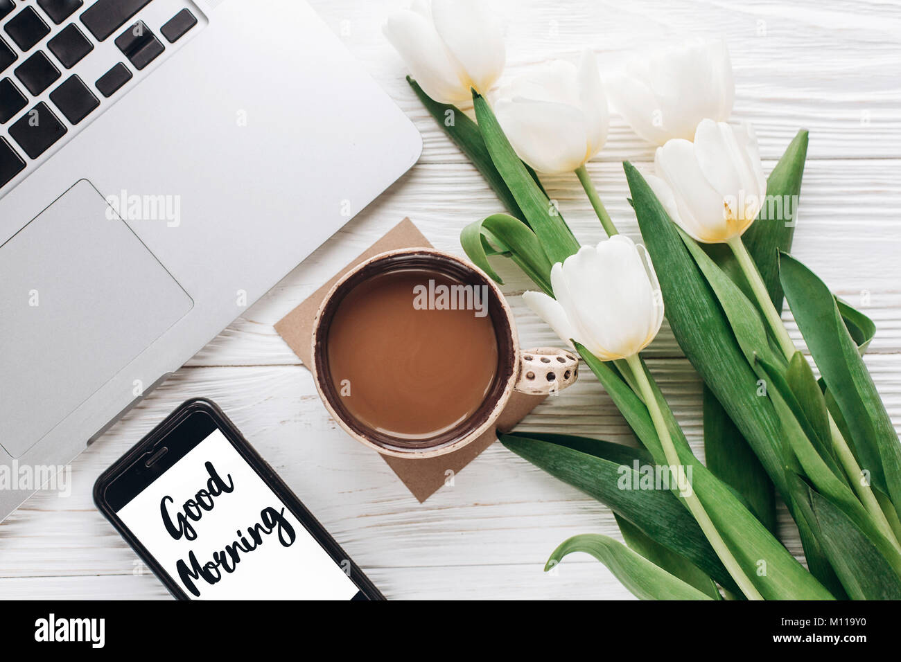 Guten Morgen text Zeichen auf Telefonbildschirm und Laptop mit Kaffee am Morgen und Tulpen auf weißen Holzmöbeln rustikal Hintergrund. stylisch Flach mit Blumen und Stockfoto