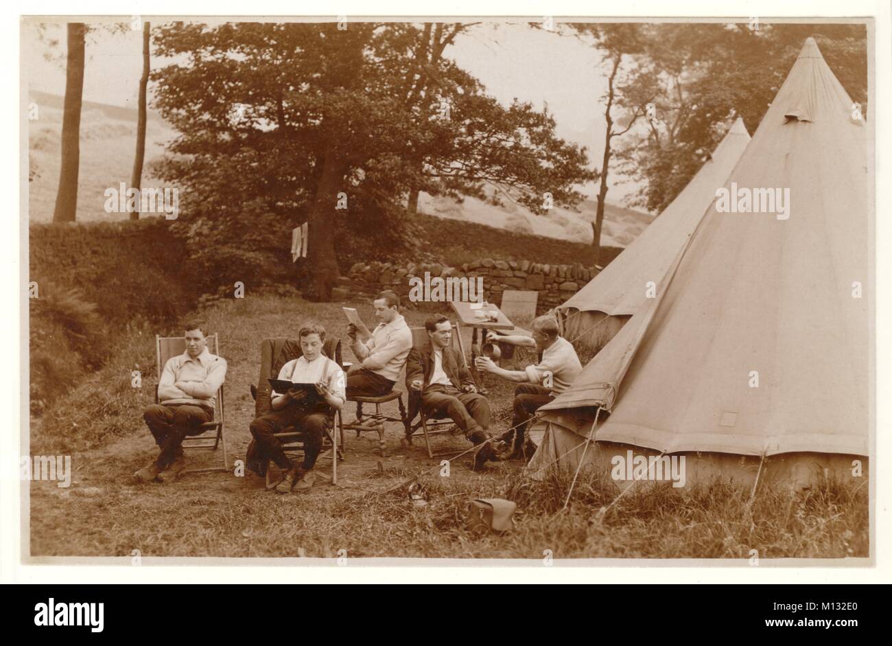 Early1900s Postkarte von jungen Männern Camping, Entspannung auf Stühlen draußen. Ein Mann liest ein Buch, einer eine Zeitung und ein anderer gießt eine Tasse Tee. Neu gemähtes Heu trocknet in Reihen von Stooks auf den Feldern dahinter. Circa Sommer 1920er, Großbritannien Stockfoto