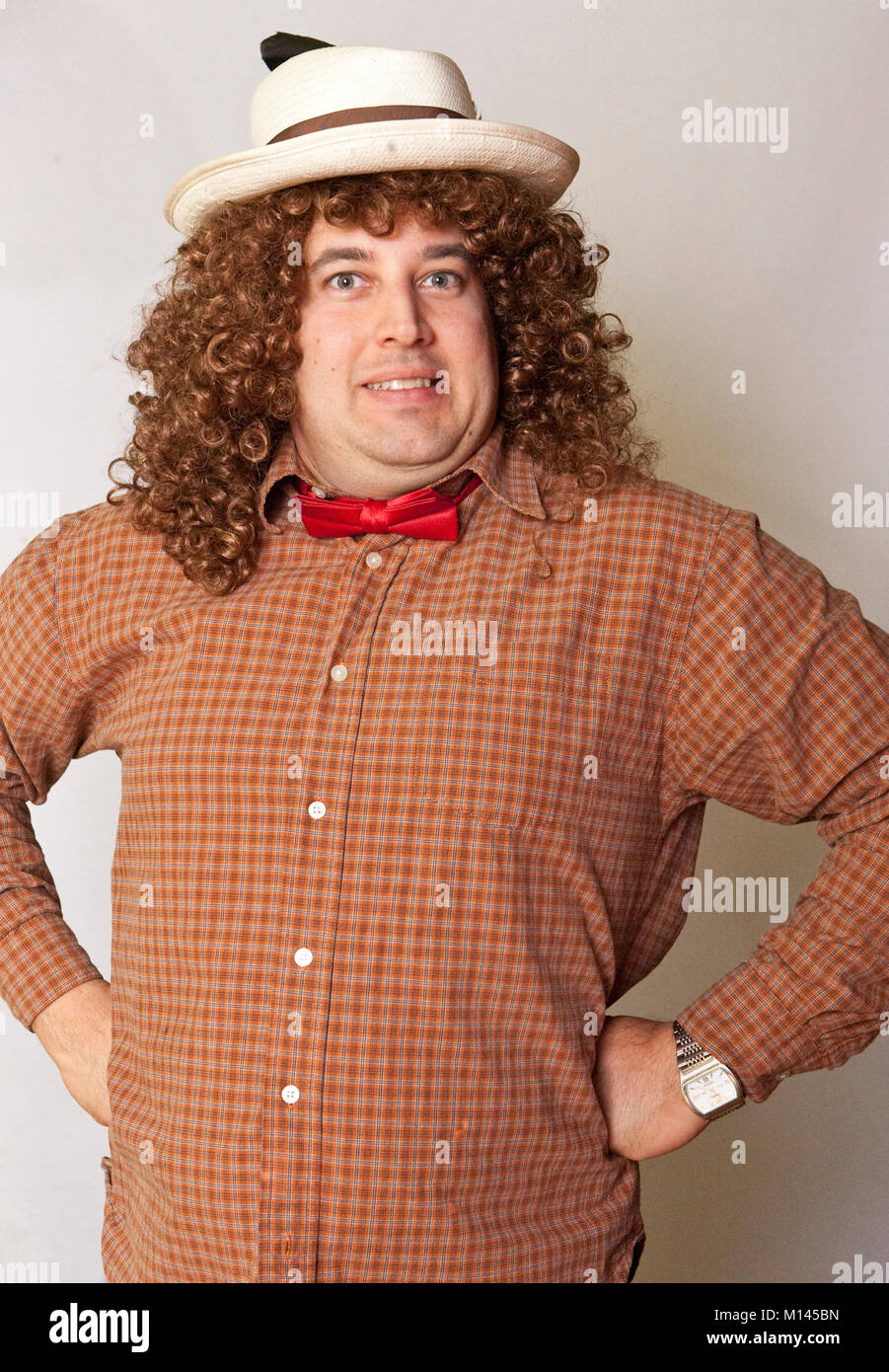 Mann mit seiner Hände auf seine Hüften, gekleidet in ein albernes Kostüm, mit einem karierten Hemd, Roter Fliege, lange lockige Haare, und einen Strohhut. Stockfoto