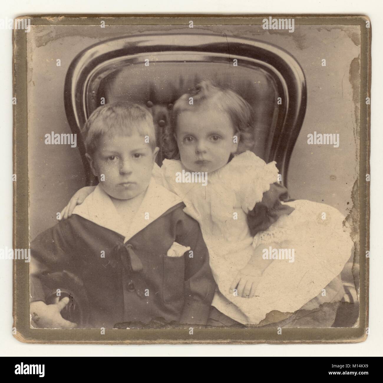 Charmantes, original viktorianisches Studioporträt eines Jungen mit seiner kleinen Schwester, Geschwistern, die in einem Sessel mit Knopfrücken sitzt. Der Junge trägt einen modischen Matrosenanzug. Ca. 1890er, Großbritannien Stockfoto