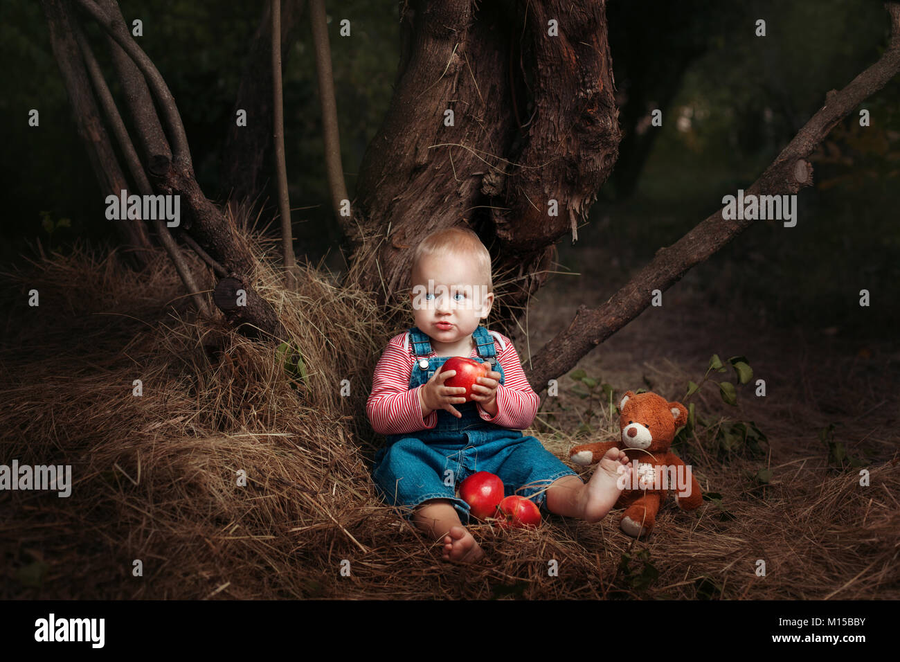 Kleines Mädchen mit einem Spielzeug Teddy saß unter einem Baum. Sie isst einen Apfel Stockfoto