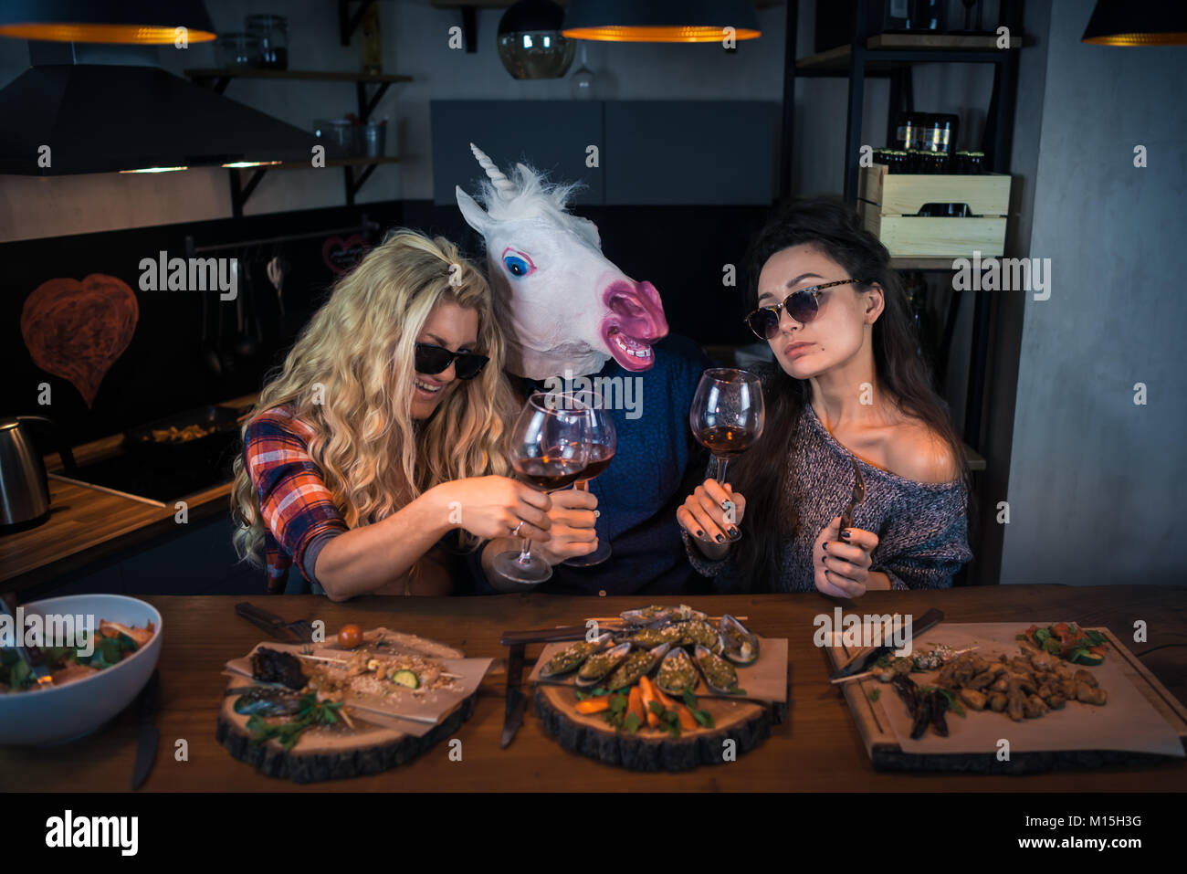Gruppe von Menschen hat das Abendessen und hält Gläser Wein. Zwei Frauen mit Einhorn genießen die Zeit zusammen. Mann in einem lustigen Gummi Maske Stockfoto