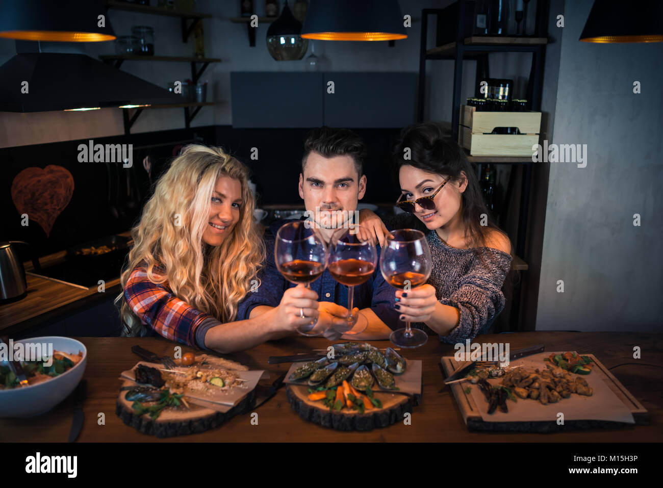 Zwei Frauen mit einem Mann lächelt und hat ein gemeinsames Abendessen. Gruppe von glücklichen Menschen in die Kamera schauen und hält Gläser Wein. Die jungen Menschen Zeit verbringen Stockfoto