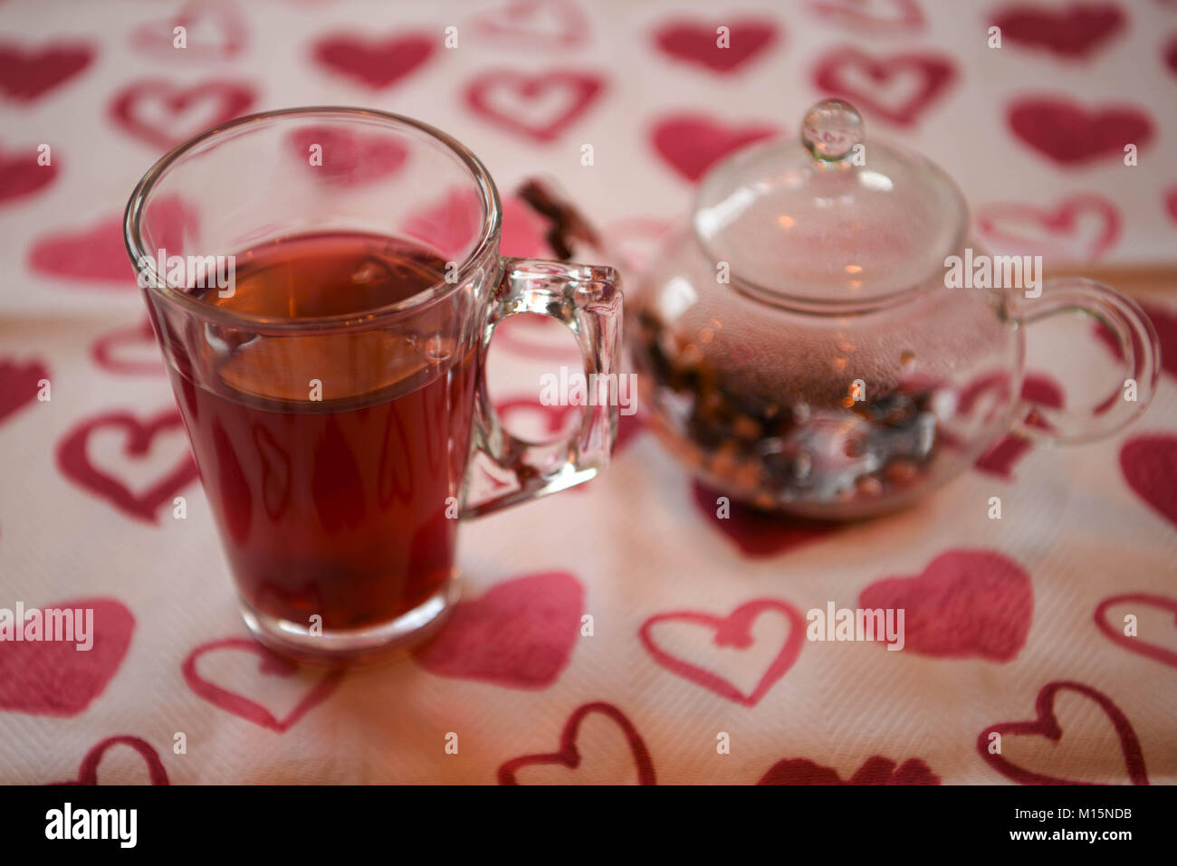 Trinken Bild eines gesunden hot Apple und Gewürz Aroma Früchtetee in einer Teekanne aus Glas mit Trinkglas und auf einem roten Liebe Herz Muster Tischdecke Stockfoto