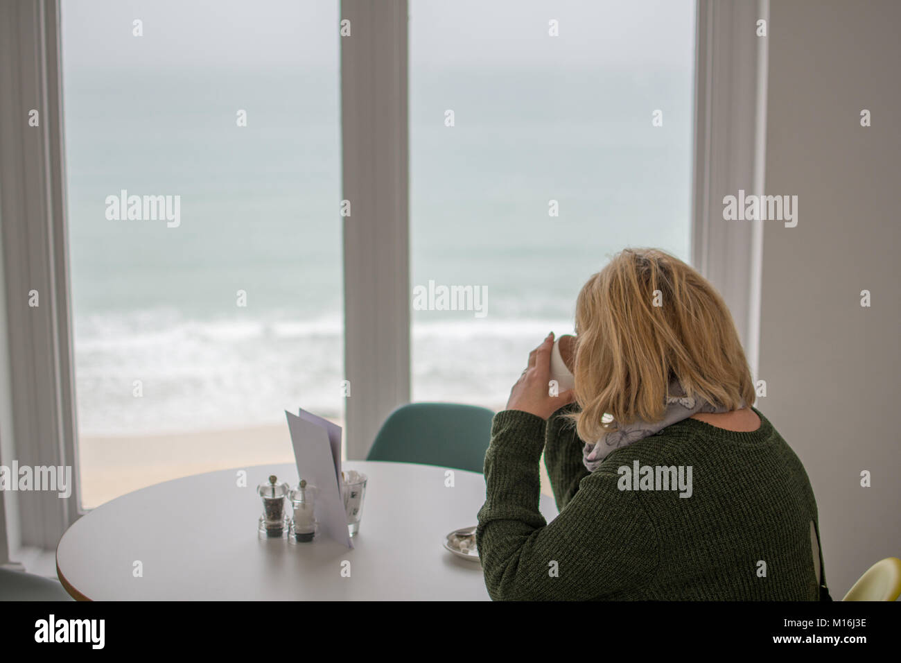 Frau trinkt Kaffee vor einem Fenster mit Blick auf den Strand und das Meer in St. Ives in Cornwall. Stockfoto