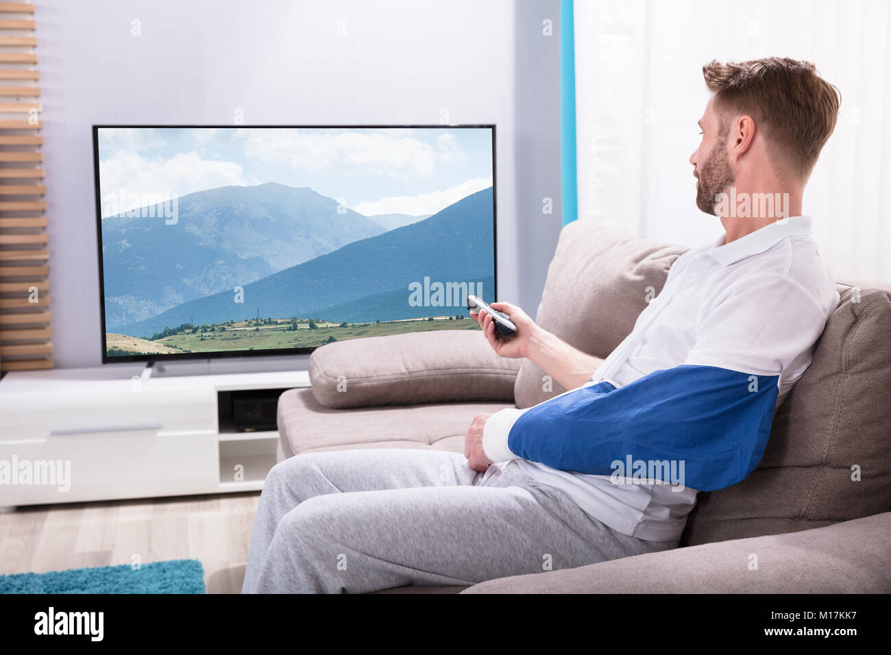 Junger Mann mit gebrochenen Hand sitzen auf dem Sofa vor dem Fernseher zu Hause Stockfoto