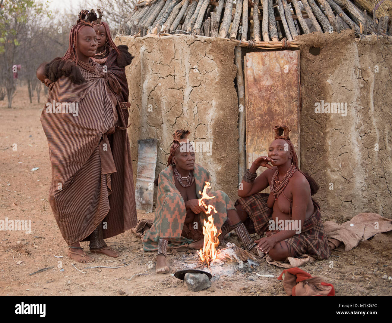 Himba Frauen sitzen am frühen Morgen vor der Lehmhütte am brennenden Feuer, Kaokoveld, Kunene, Namibia Stockfoto