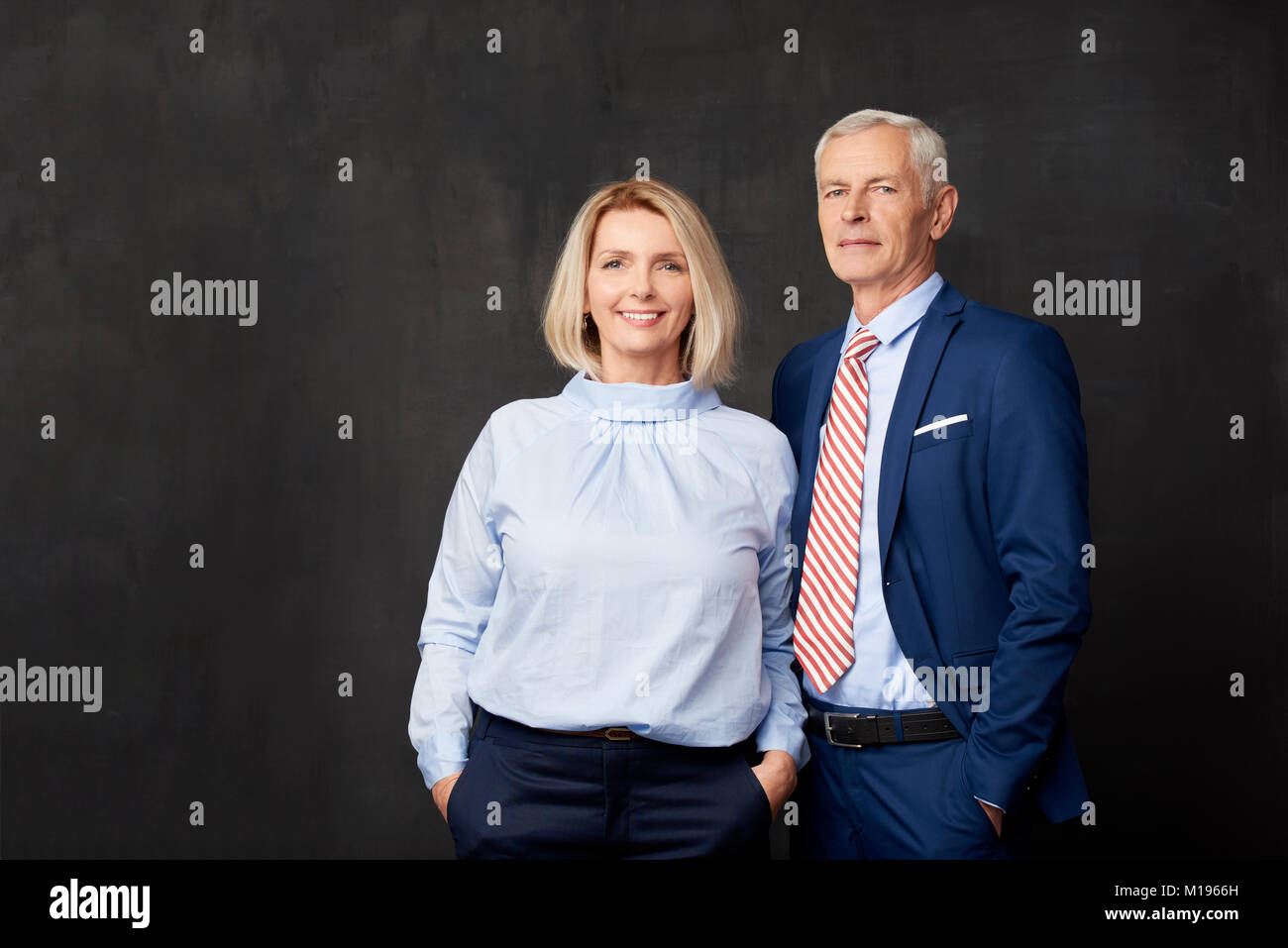 Senior Paar stehen gemeinsam auf dunklem Hintergrund. Schöne blonde Frau an der Kamera schauen und lächeln, während eleganten alten Mann neben ihr. Stockfoto