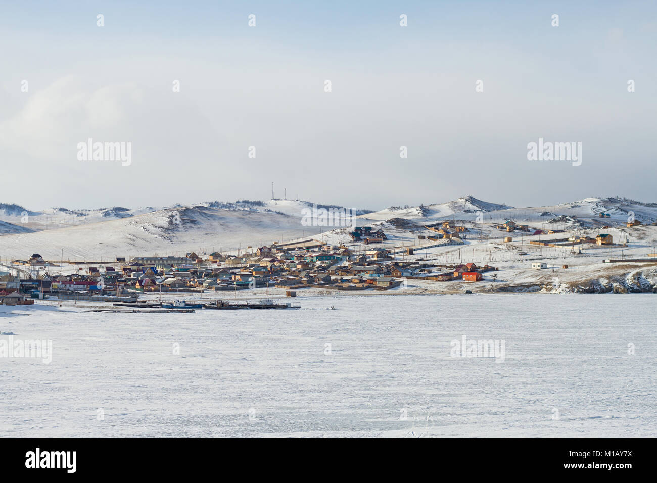 Dorf Landschaft mit Häusern im Winter am Ufer des Baikalsees, die Schiffe auf dem Eis auf einem schneebedeckten Bergen im Hintergrund an einem bewölkten Tag. Stockfoto