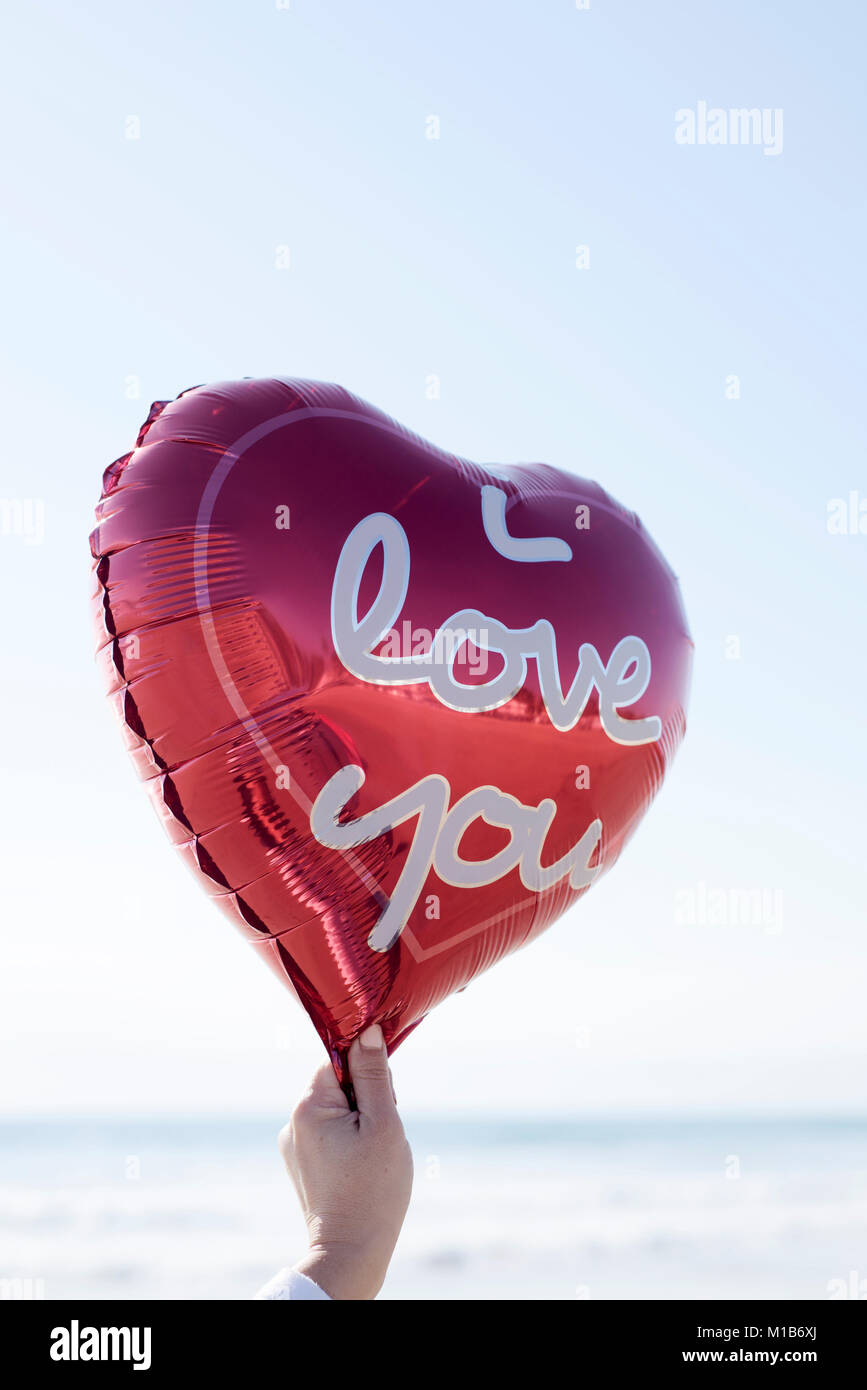 Nahaufnahme der Hand einer jungen Frau mit einem roten herzförmigen Luftballon mit dem Text "Ich liebe Dich in Es geschrieben, gegen den Himmel und das Meer in der Bac Stockfoto