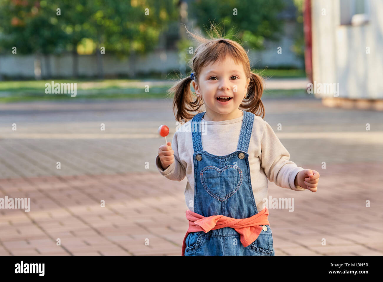 Kleine hübsche Mädchen essen Süßigkeiten im Sommer Park. Adorable kleine Mädchen in Jeans Overalls lächelnd. Porträt eines Kindes 2 Jahre alt, Soft Focus. Stockfoto