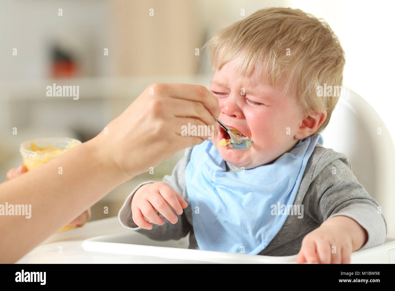 Nahaufnahme der Mutter Hand Feeding seinen Sohn, der weinend sitzt auf einem hohen Stuhl Stockfoto