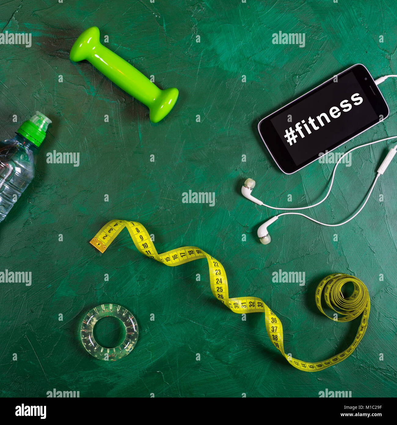 Hantel, Wasserflasche, Smartphone, Kopfhörer und Maßband auf grünem Hintergrund. Hashtag Fitness auf dem Bildschirm des Smartphones Stockfoto