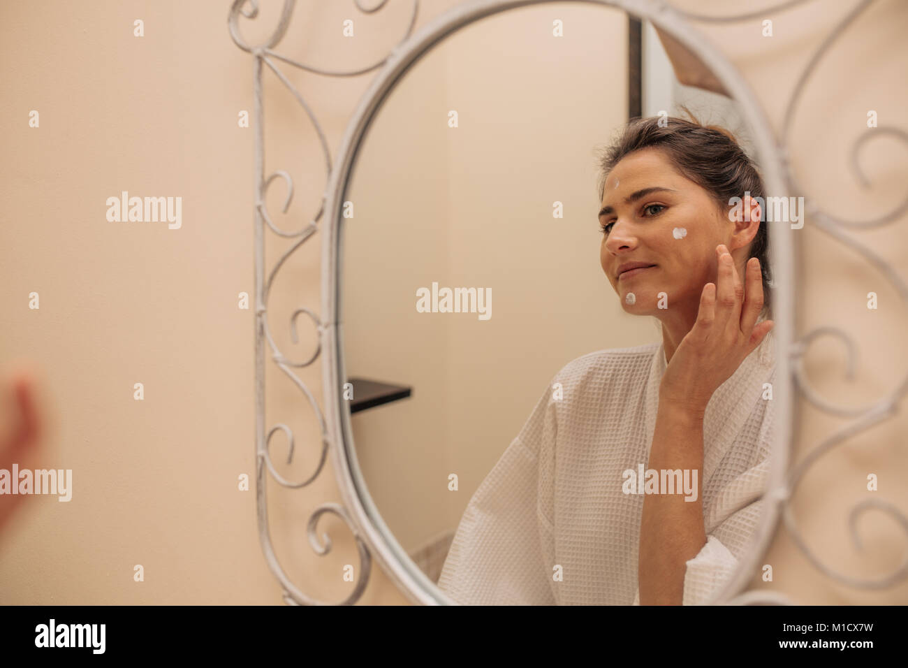 Frau Anwendung Feuchtigkeits-creme auf Gesicht, während im Bad. Reflexion der weiblichen Blick in den Spiegel und Kosmetik Lotion auf ihrem Gesicht. Stockfoto