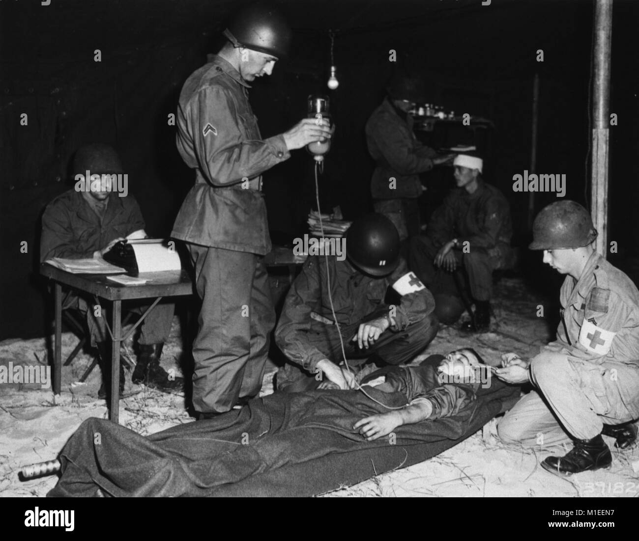 Die verwundeten Soldaten liegt auf einer Bahre in einem Zelt, 7. Dezember 1951. Drei Soldaten dazu neigen, den Patienten: Eine kniet und nimmt seinen Puls, ein anderer hockt und macht sich Notizen, und ein dritter steht und hält ein Glas IV mit einer Flüssigkeit, die angezeigt wird, ein Produkt zu sein, vielleicht Plasma. Hinter ihnen eine andere verwundete Soldaten gepflegt wird, während ein anderer an einem Tisch und Typen sitzt, Washington DC. Mit freundlicher Genehmigung der nationalen Bibliothek für Medizin. () Stockfoto
