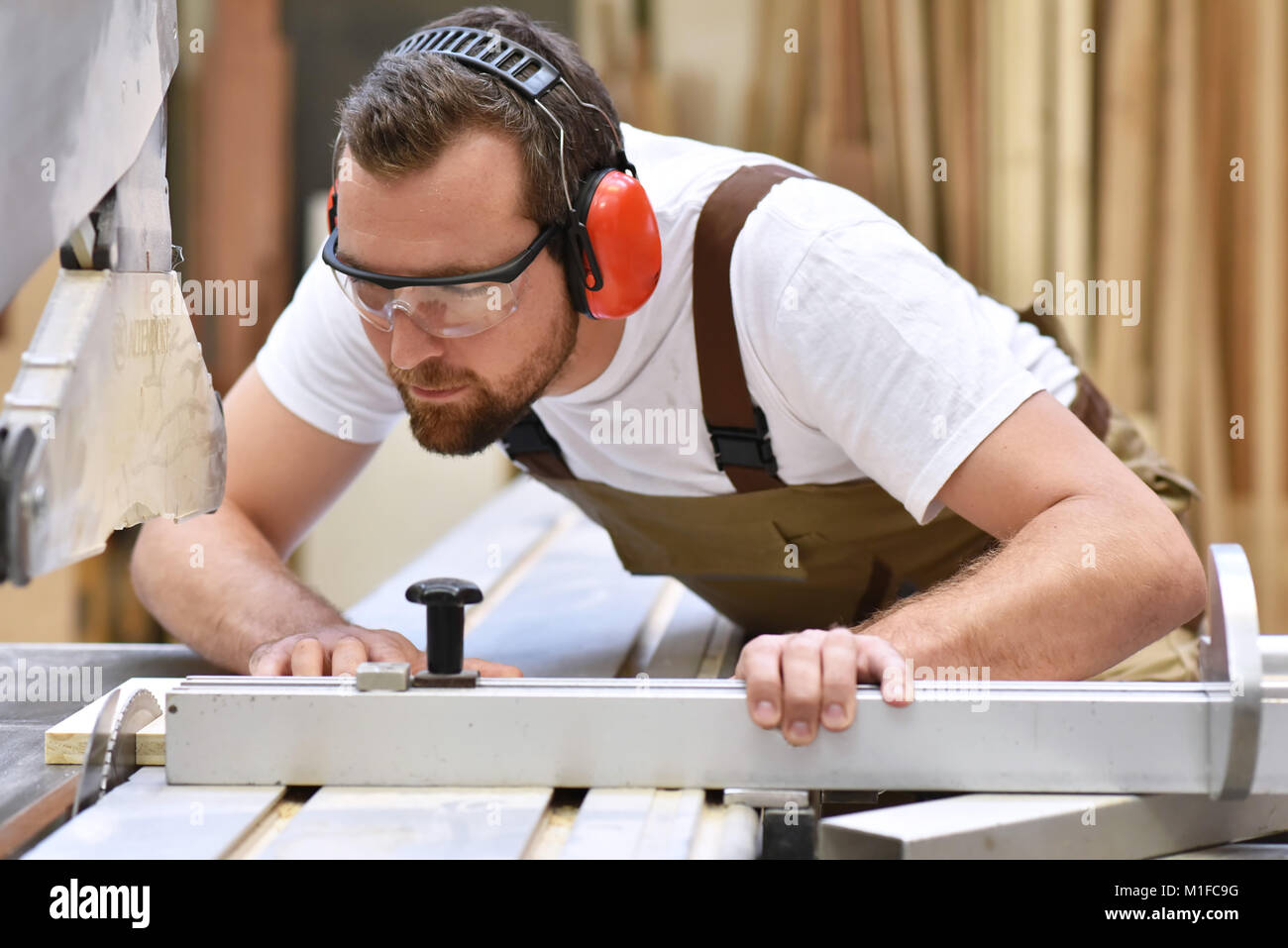 Junge Tischler in Arbeitskleidung arbeitet in der Tischlerei auf einer Schleifmaschine - Arbeitskleidung mit Schutzbrille und Gehörschutz Stockfoto