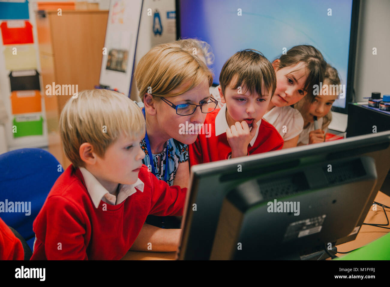 Grundschüler stehen rund um ihren Lehrer am Computer im Klassenzimmer. Sie sind alle Konzentration auf etwas auf dem Bildschirm. Stockfoto