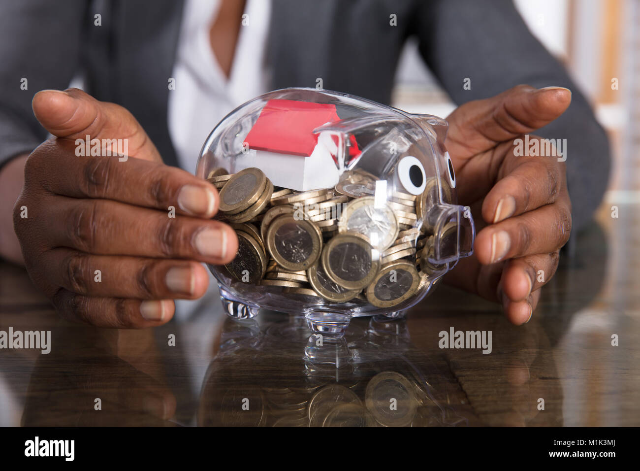 Nahaufnahme einer menschlichen Hand Schutz Sparschwein mit Haus Modell und Münzen am Schreibtisch Stockfoto